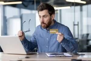 a guy holding a credit card surprised at the credit card amount showing on his laptop